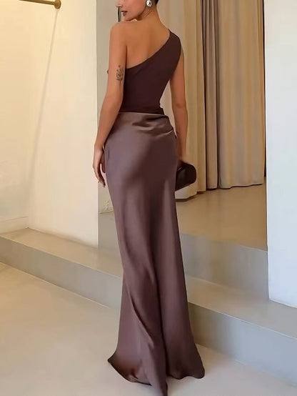 Blue - A woman wears a floor-length, one-shoulder maxi dress in a deep chocolate brown. The dress features a matte, draped bodice with an asymmetric waistline that flows into a lustrous, liquid-satin skirt.