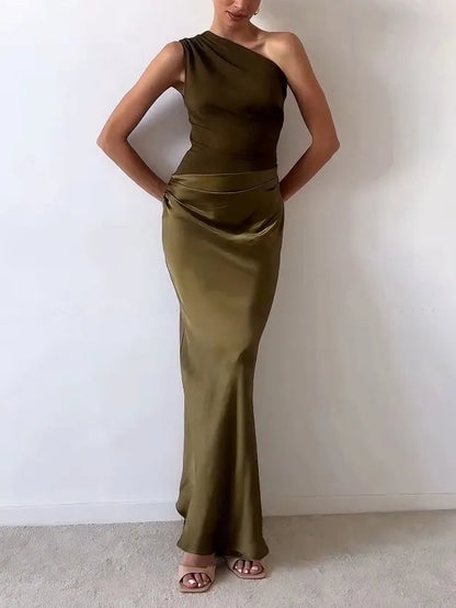 Blue - A woman wears a floor-length, one-shoulder maxi dress in a deep chocolate brown. The dress features a matte, draped bodice with an asymmetric waistline that flows into a lustrous, liquid-satin skirt.