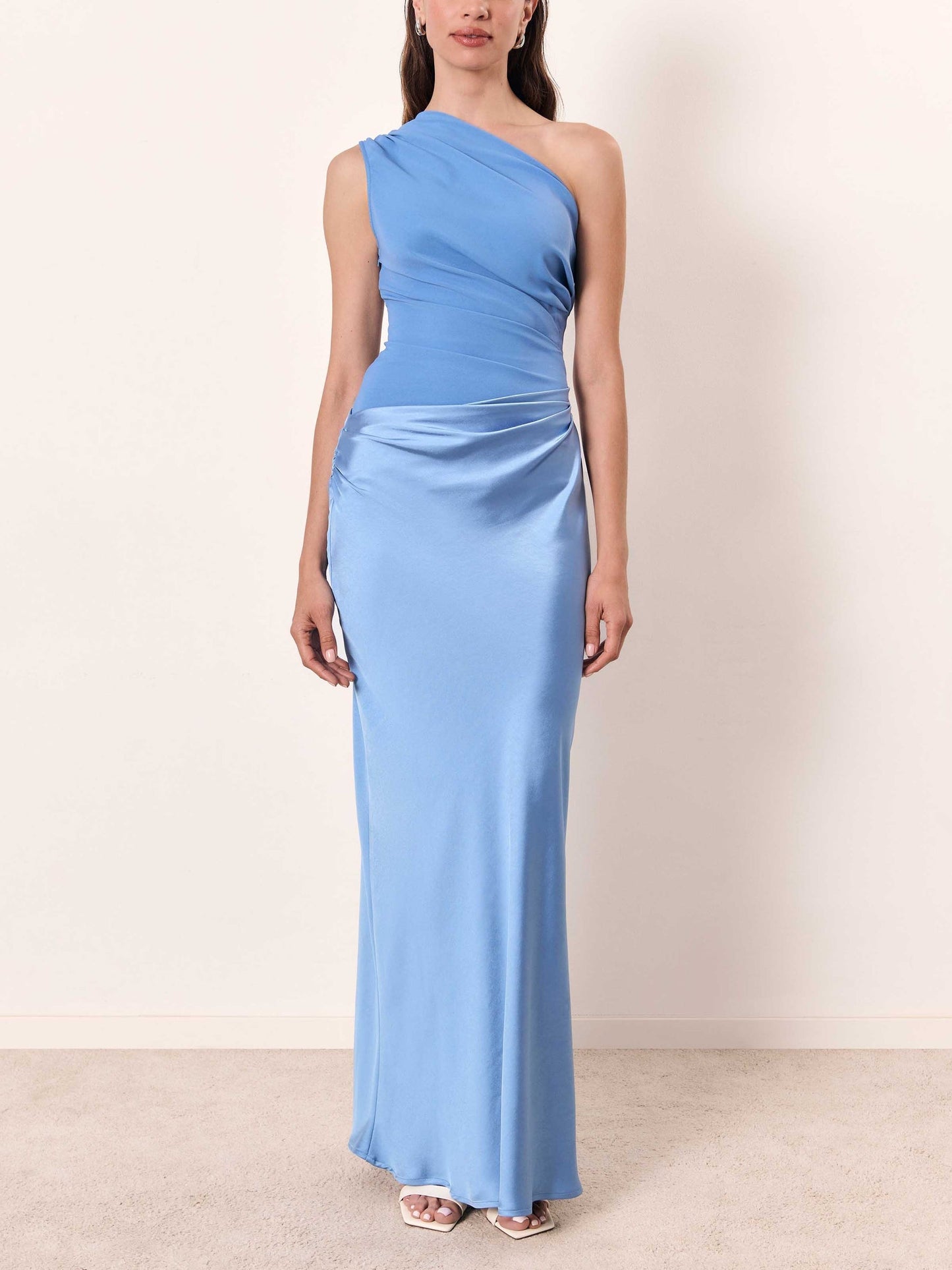 Blue - A woman wears a floor-length, one-shoulder maxi dress in a deep chocolate brown. The dress features a matte, draped bodice with an asymmetric waistline that flows into a lustrous, liquid-satin skirt.