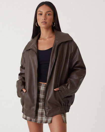 Brown - A woman wearing an oversized dark brown faux leather bomber jacket with a wide collar, styled over a black top and plaid mini skirt.