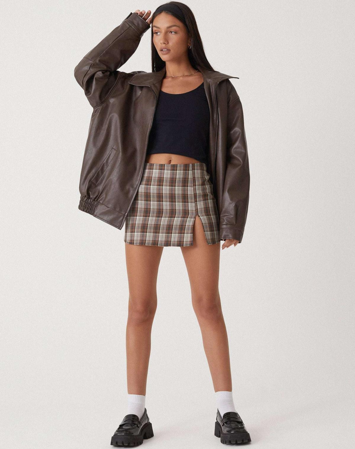 Brown - A woman wearing an oversized dark brown faux leather bomber jacket with a wide collar, styled over a black top and plaid mini skirt.