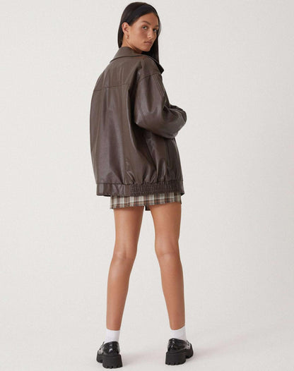 Brown - A woman wearing an oversized dark brown faux leather bomber jacket with a wide collar, styled over a black top and plaid mini skirt.