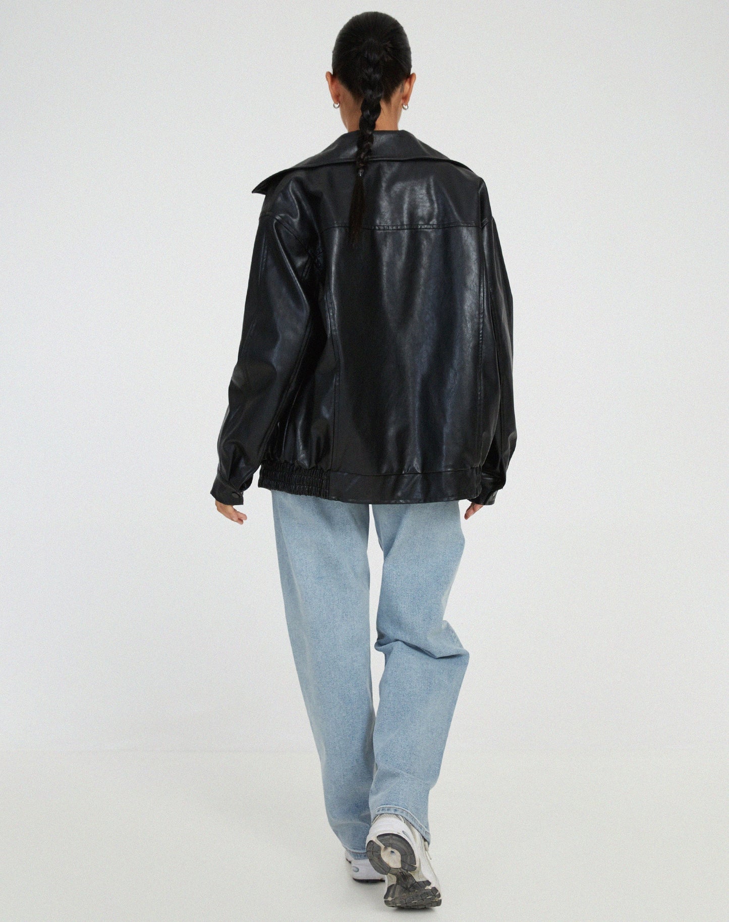 Black - A woman wearing an oversized dark brown faux leather bomber jacket with a wide collar, styled over a black top and plaid mini skirt.