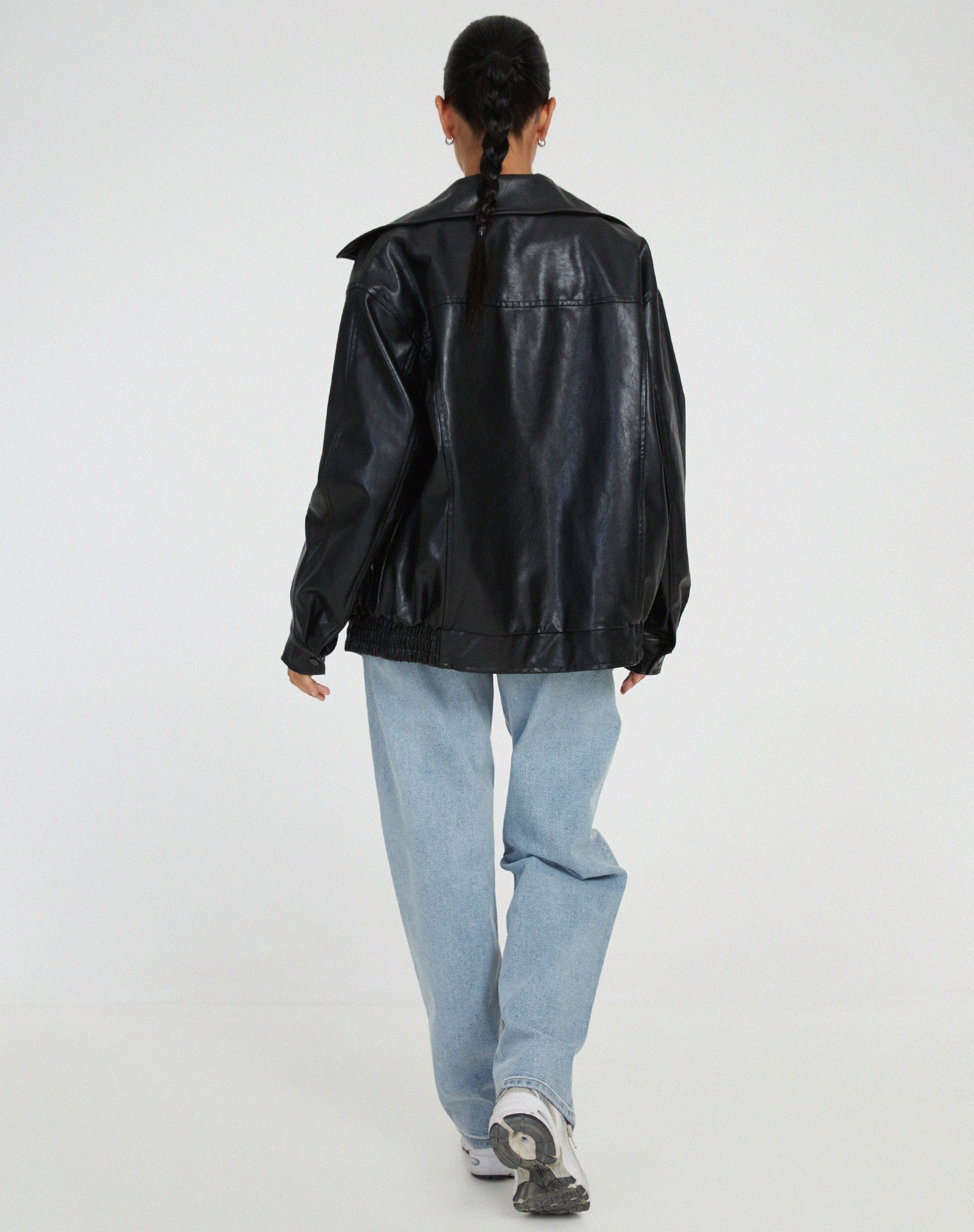 Black - A woman wearing an oversized dark brown faux leather bomber jacket with a wide collar, styled over a black top and plaid mini skirt.