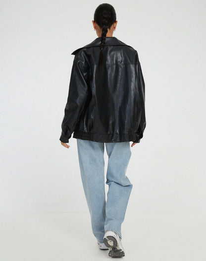 Black - A woman wearing an oversized dark brown faux leather bomber jacket with a wide collar, styled over a black top and plaid mini skirt.
