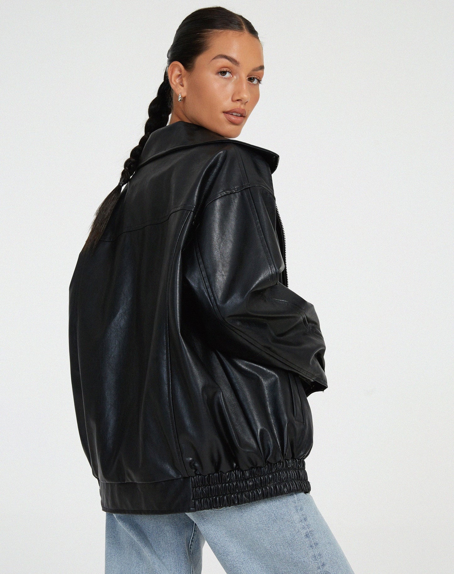 Black - A woman wearing an oversized dark brown faux leather bomber jacket with a wide collar, styled over a black top and plaid mini skirt.