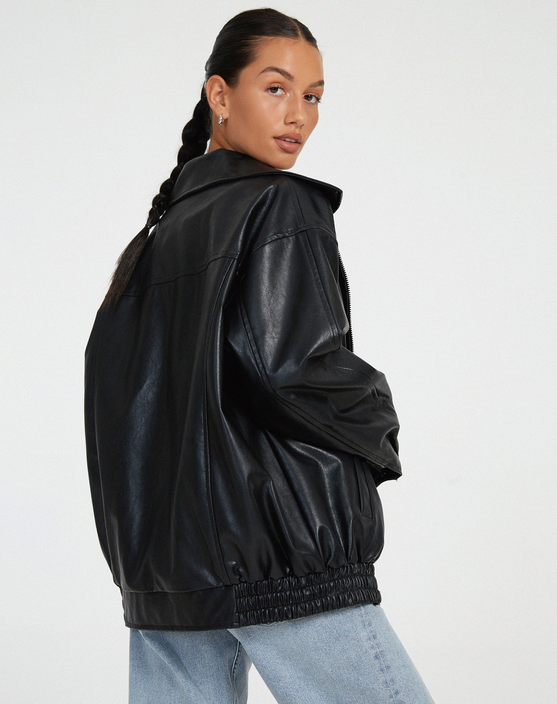 Black - A woman wearing an oversized dark brown faux leather bomber jacket with a wide collar, styled over a black top and plaid mini skirt.