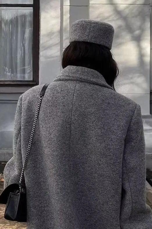 Grey - A woman wearing an oversized, double-breasted maxi coat in textured heather grey wool, featuring wide notched lapels and flap pockets, paired with a matching grey pillbox hat.