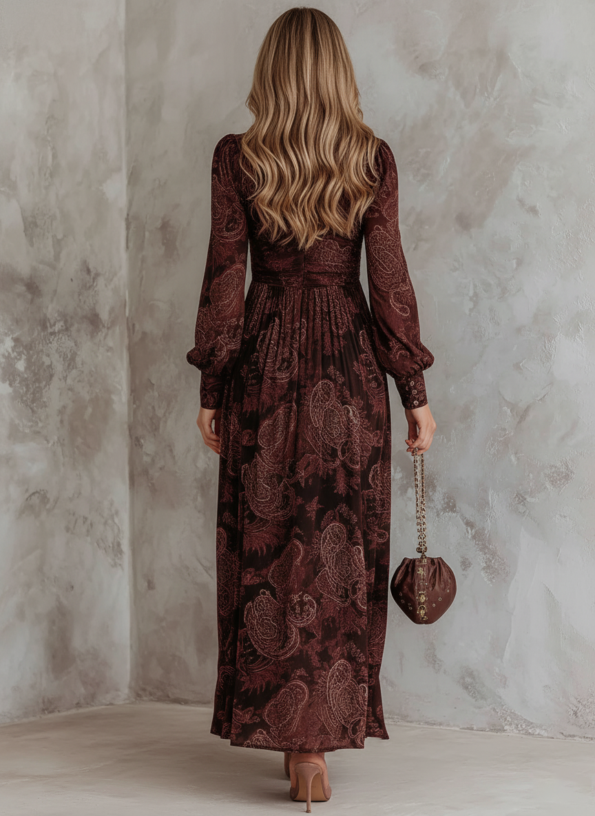 2XL - A woman wearing a long-sleeved, brown paisley print maxi dress with a zippered V-neckline, ruched bodice, and flowing skirt, holding a small brown satin handbag.