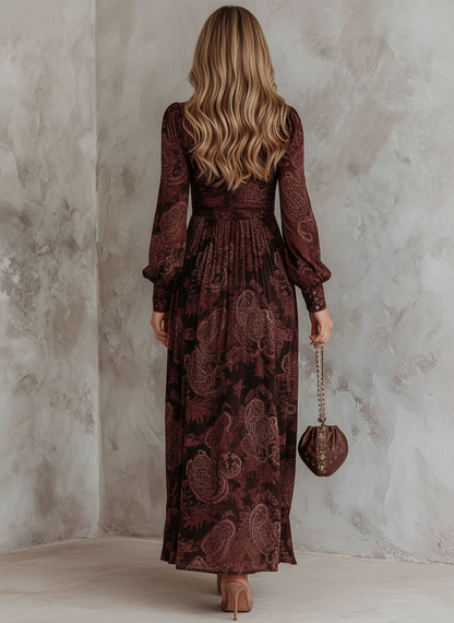 2XL - A woman wearing a long-sleeved, brown paisley print maxi dress with a zippered V-neckline, ruched bodice, and flowing skirt, holding a small brown satin handbag.