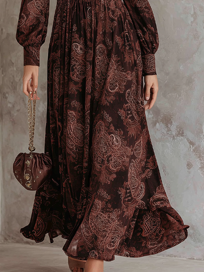 2XL - A woman wearing a long-sleeved, brown paisley print maxi dress with a zippered V-neckline, ruched bodice, and flowing skirt, holding a small brown satin handbag.