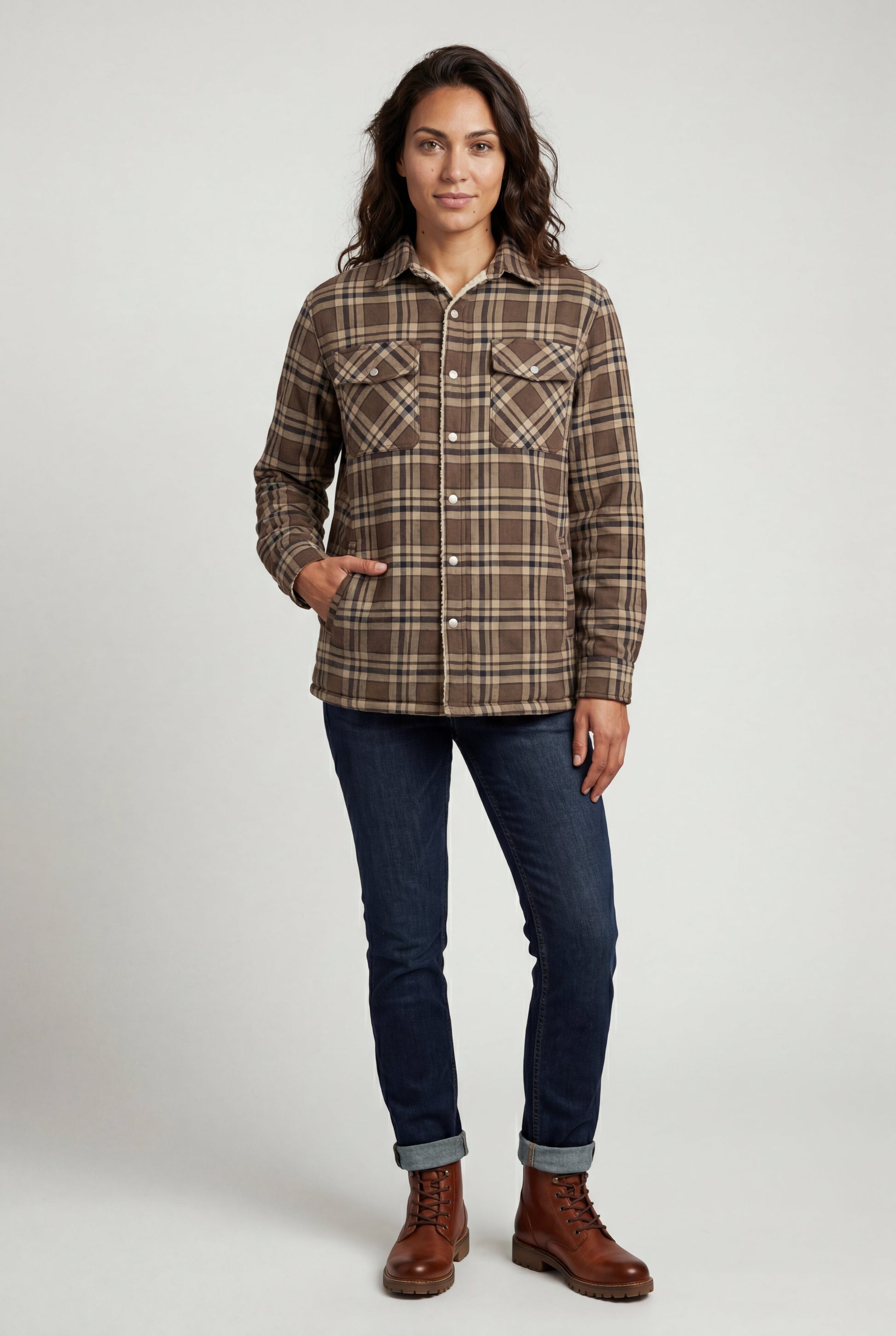 Khaki - A woman wearing a brown plaid check shirt jacket with a cream-coloured sherpa lining, styled with dark blue slim-fit jeans and brown leather lace-up boots against a plain grey background.