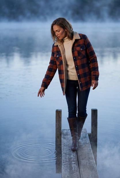 Green - A woman wearing a brown plaid check shirt jacket with a cream-coloured sherpa lining, styled with dark blue slim-fit jeans and brown leather lace-up boots against a plain grey background.