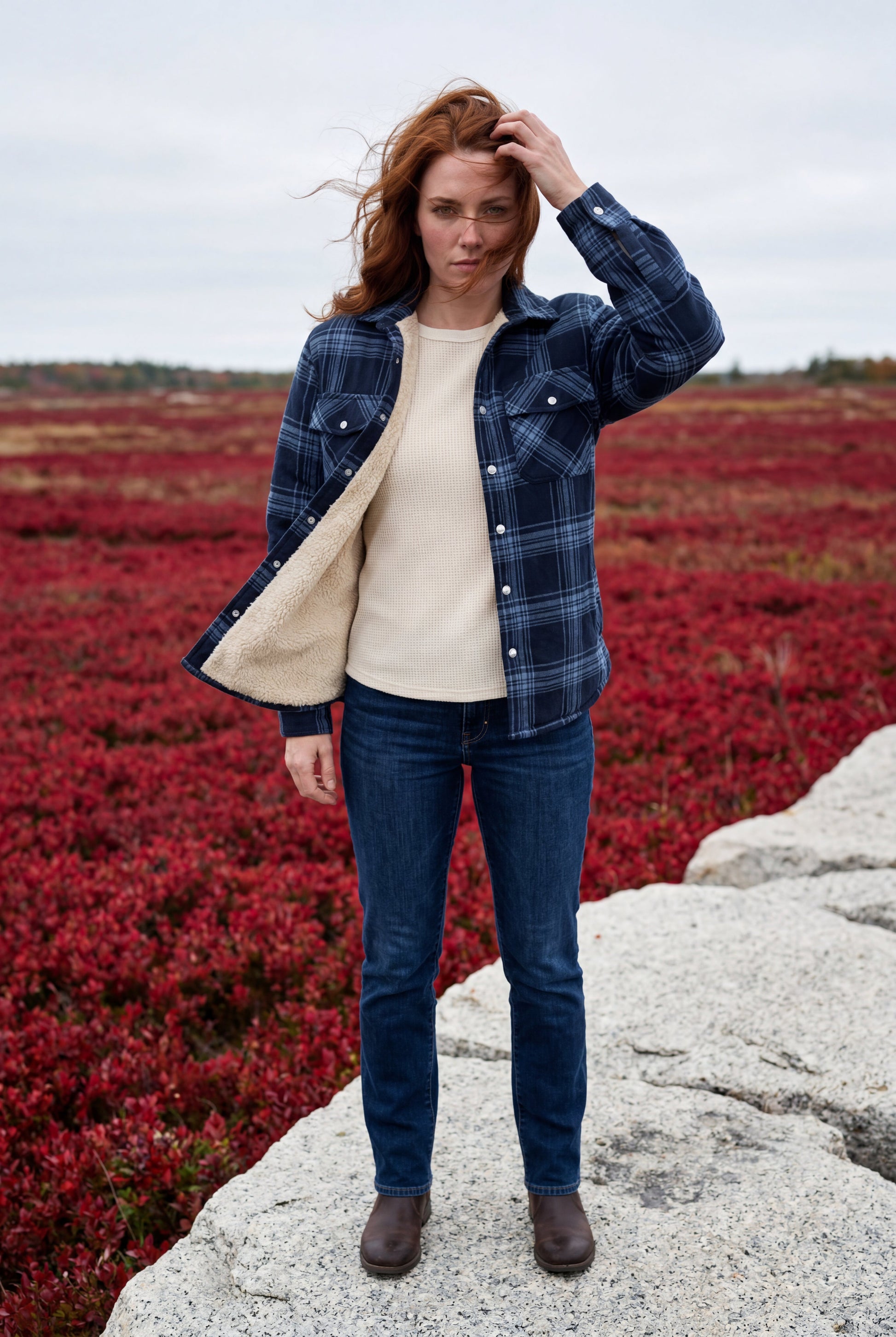 Denim Blue - A woman wearing a brown plaid check shirt jacket with a cream-coloured sherpa lining, styled with dark blue slim-fit jeans and brown leather lace-up boots against a plain grey background.