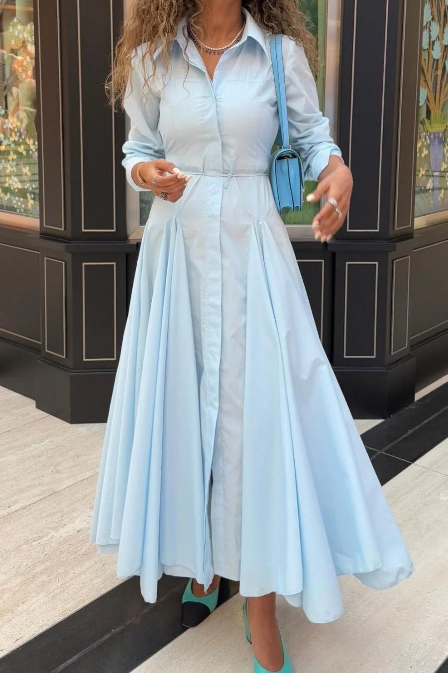 Lake Blue - Woman wearing a light blue, long-sleeved midi shirt dress with a collared neckline, button-front, and a voluminous pleated skirt.