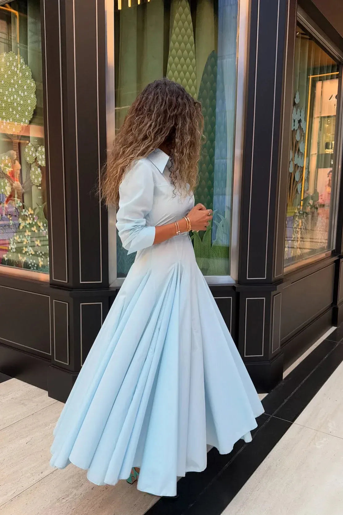 Lake Blue - Woman wearing a light blue, long-sleeved midi shirt dress with a collared neckline, button-front, and a voluminous pleated skirt.