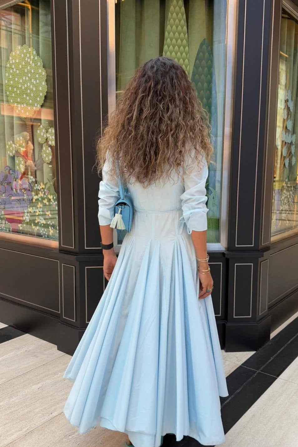 Lake Blue - Woman wearing a light blue, long-sleeved midi shirt dress with a collared neckline, button-front, and a voluminous pleated skirt.