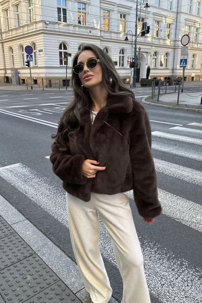 Brown - A woman wearing a dark brown plush faux fur cropped jacket with a wide collar, styled with a brown mini skirt, black knee-high boots, and holding a black leather handbag against a stone building background.