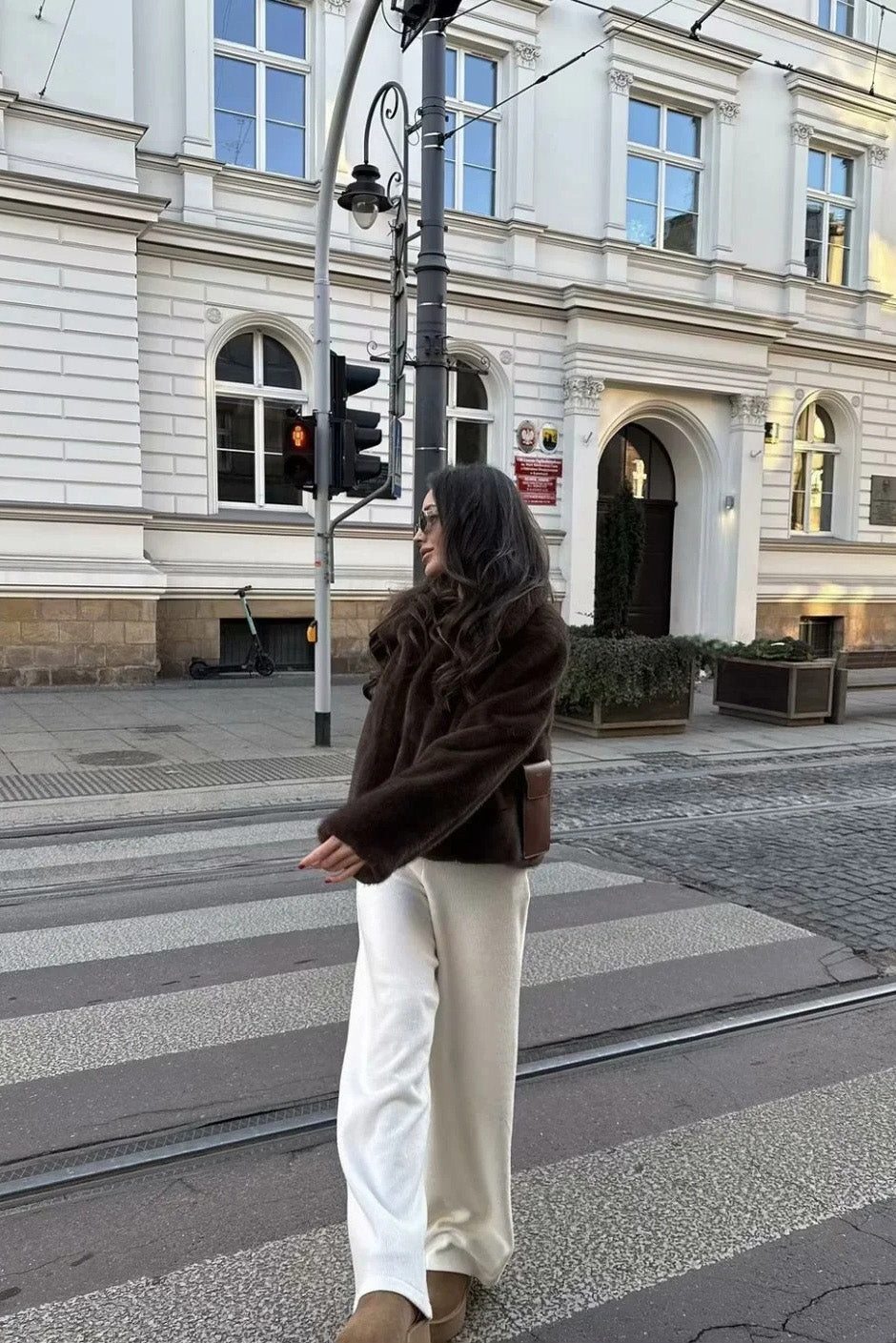 A woman wearing a dark brown plush faux fur cropped jacket with a wide collar, styled with a brown mini skirt, black knee-high boots, and holding a black leather handbag against a stone building background.