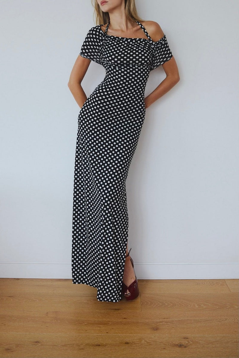 Black - A woman wearing a black and white polka dot maxi dress with halter straps and draped off-the-shoulder sleeves, posing against a white wall.