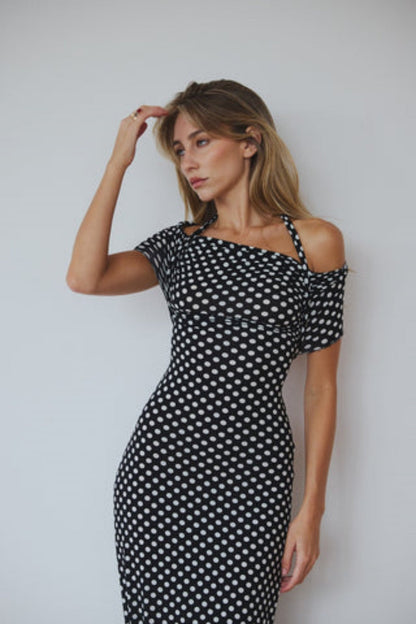 Black - A woman wearing a black and white polka dot maxi dress with halter straps and draped off-the-shoulder sleeves, posing against a white wall.
