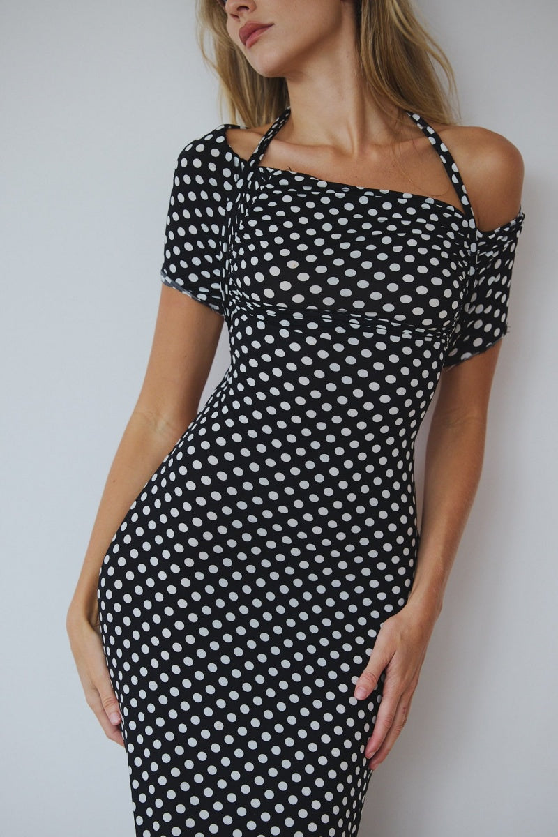 Black - A woman wearing a black and white polka dot maxi dress with halter straps and draped off-the-shoulder sleeves, posing against a white wall.