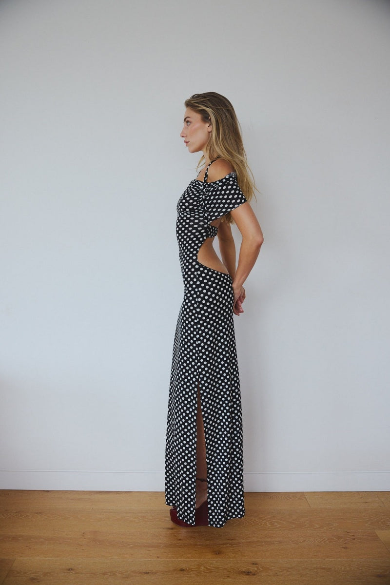 Black - A woman wearing a black and white polka dot maxi dress with halter straps and draped off-the-shoulder sleeves, posing against a white wall.