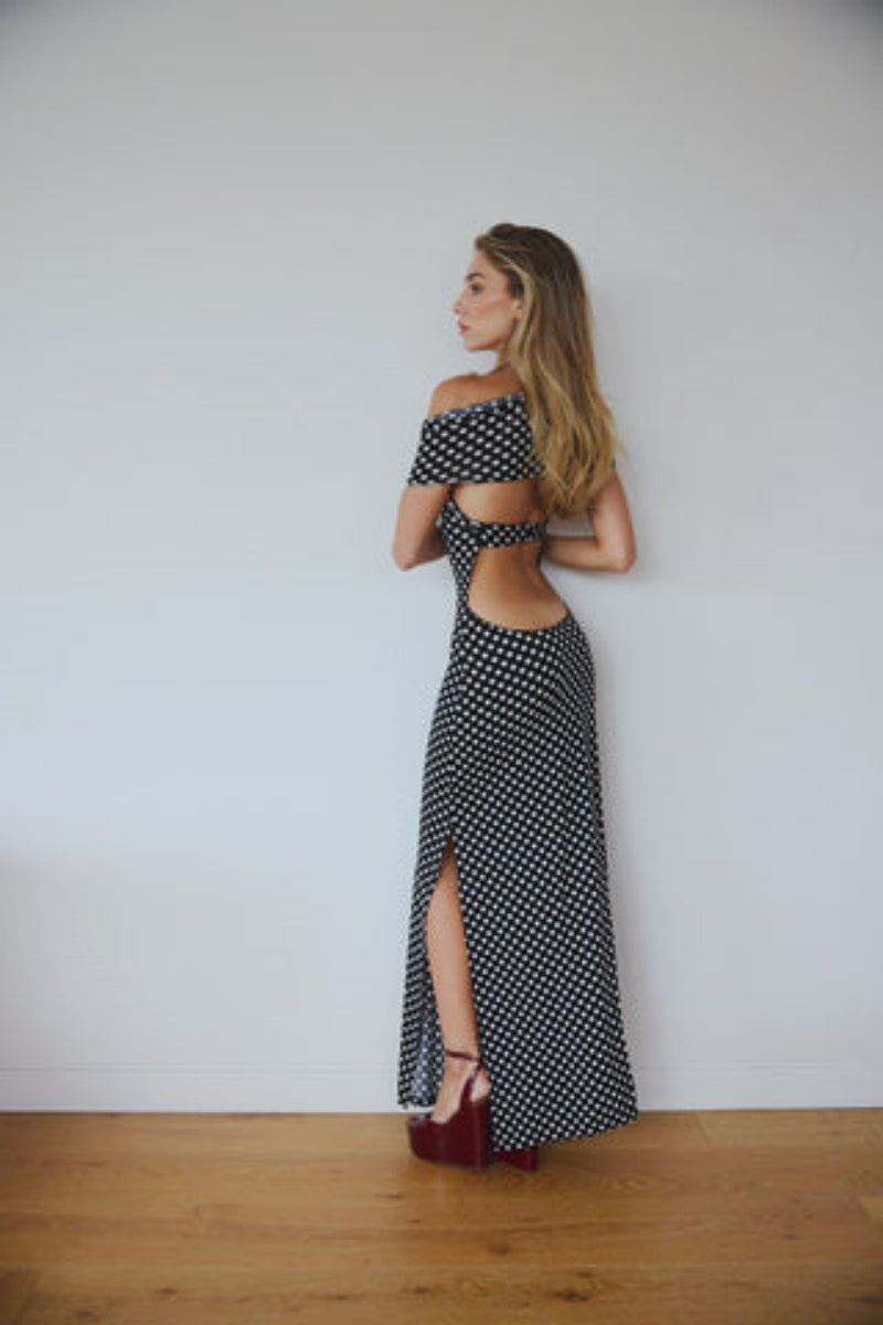 Black - A woman wearing a black and white polka dot maxi dress with halter straps and draped off-the-shoulder sleeves, posing against a white wall.