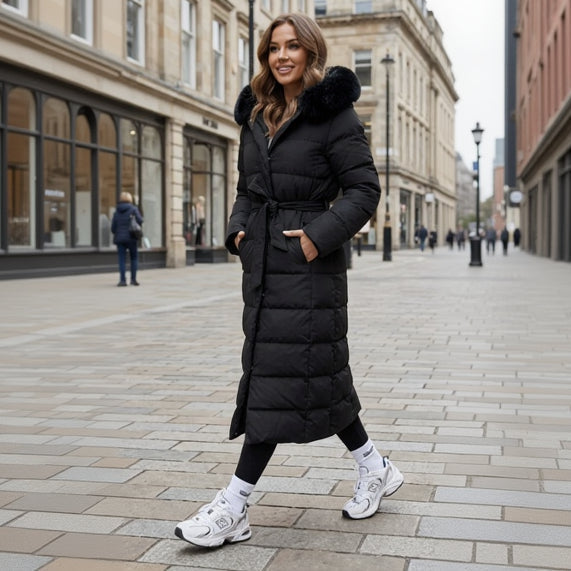 Black - A woman wearing a longline black quilted puffer coat with a plush black fur-trimmed hood and a self-tie belted waist, set against a clean white background.