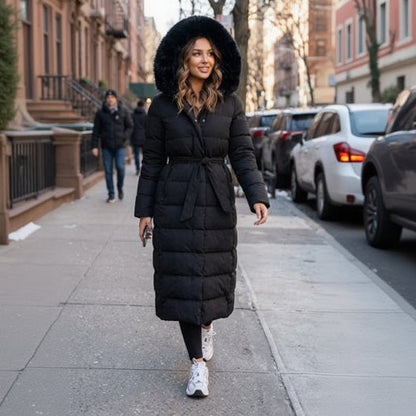 Black - A woman wearing a longline black quilted puffer coat with a plush black fur-trimmed hood and a self-tie belted waist, set against a clean white background.