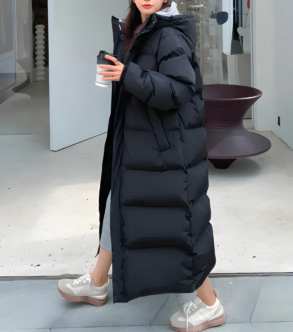 Black - A woman walking while wearing a longline, matte black quilted puffer coat with a hood, styled over a light grey outfit and chunky white sneakers.