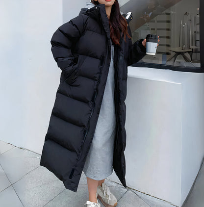 Black - A woman walking while wearing a longline, matte black quilted puffer coat with a hood, styled over a light grey outfit and chunky white sneakers.