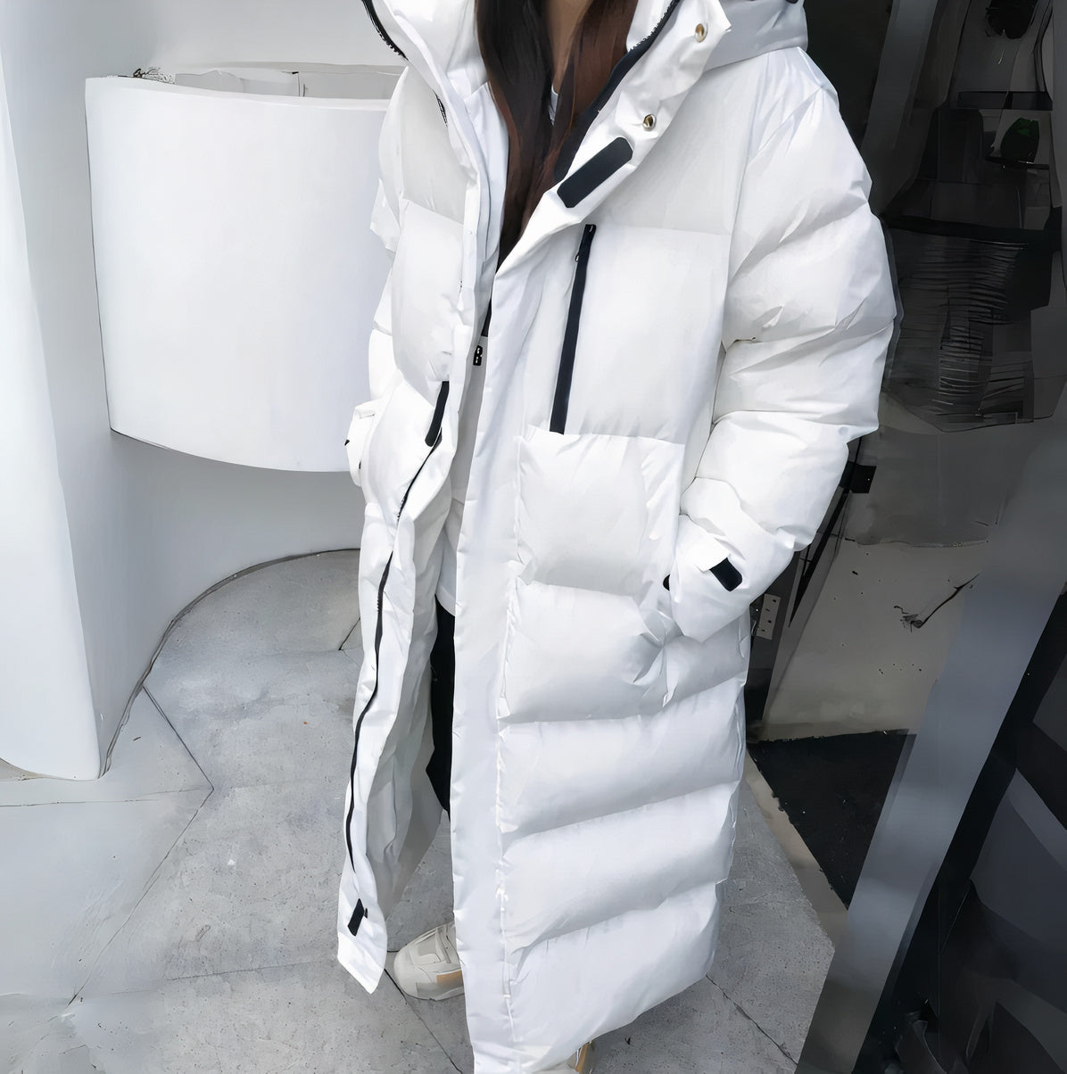 Black - A woman walking while wearing a longline, matte black quilted puffer coat with a hood, styled over a light grey outfit and chunky white sneakers.
