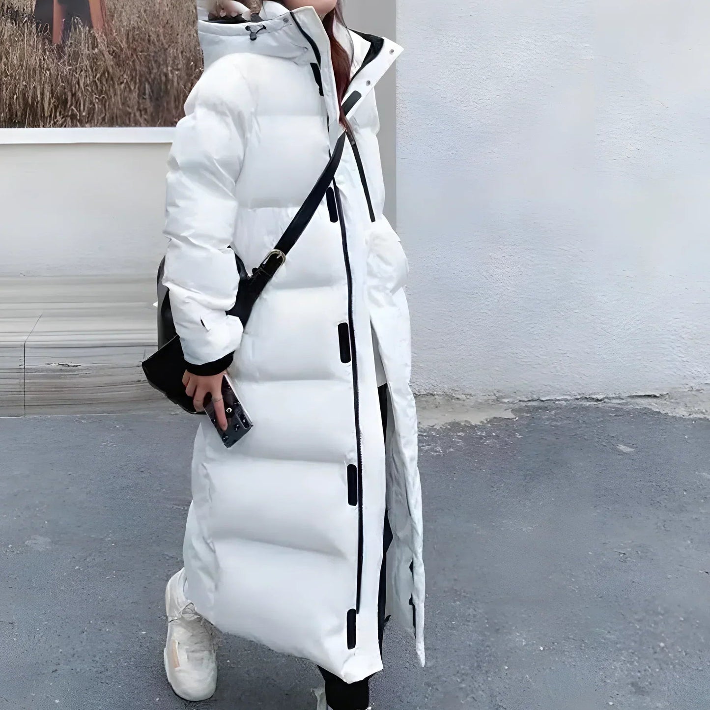 White - A woman walking while wearing a longline, matte black quilted puffer coat with a hood, styled over a light grey outfit and chunky white sneakers.