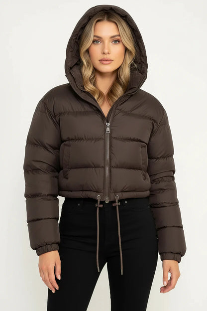 Brown - A woman wearing a dark brown cropped puffer jacket with an integrated hood, featuring horizontal quilting, a front zip closure, and adjustable drawstring toggles at the hem, paired with black high-waisted jeans.