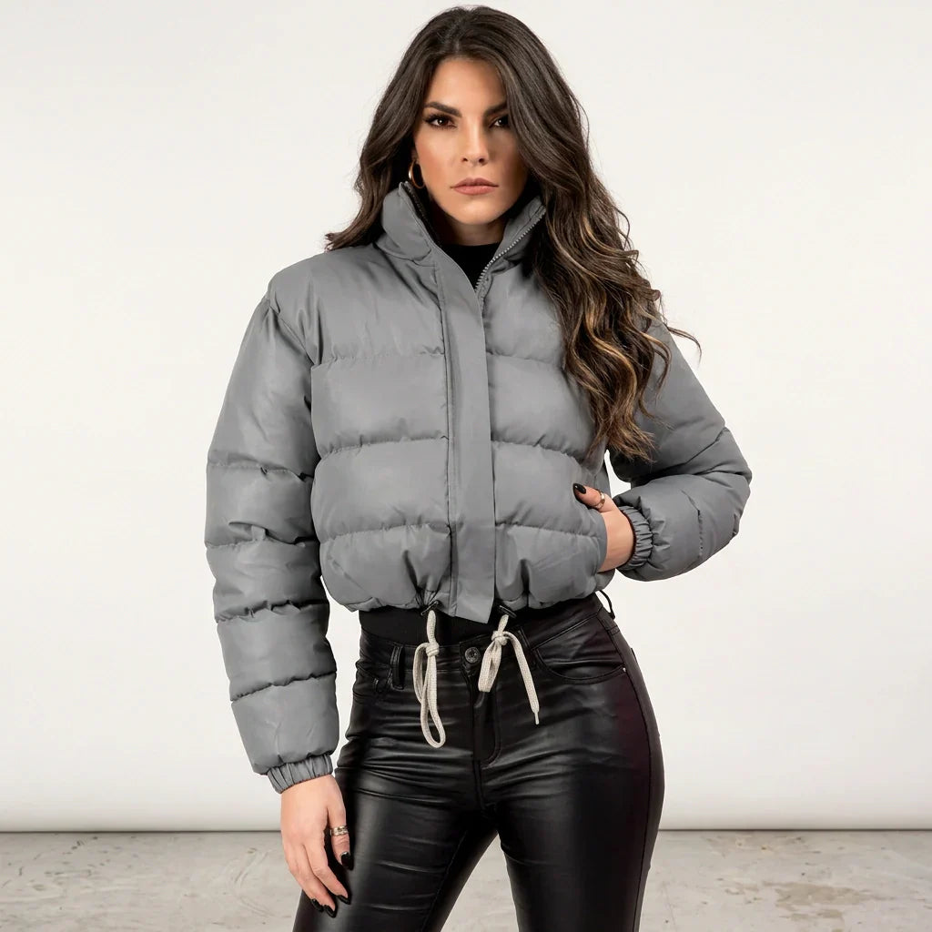 Dark Grey - A woman wearing a dark brown cropped puffer jacket with an integrated hood, featuring horizontal quilting, a front zip closure, and adjustable drawstring toggles at the hem, paired with black high-waisted jeans.