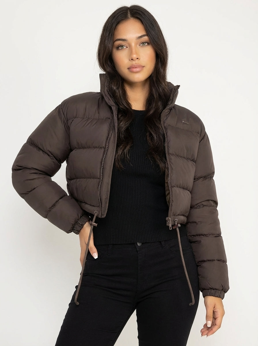 Brown - A woman wearing a dark brown cropped puffer jacket with an integrated hood, featuring horizontal quilting, a front zip closure, and adjustable drawstring toggles at the hem, paired with black high-waisted jeans.