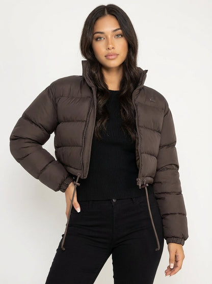 Brown - A woman wearing a dark brown cropped puffer jacket with an integrated hood, featuring horizontal quilting, a front zip closure, and adjustable drawstring toggles at the hem, paired with black high-waisted jeans.
