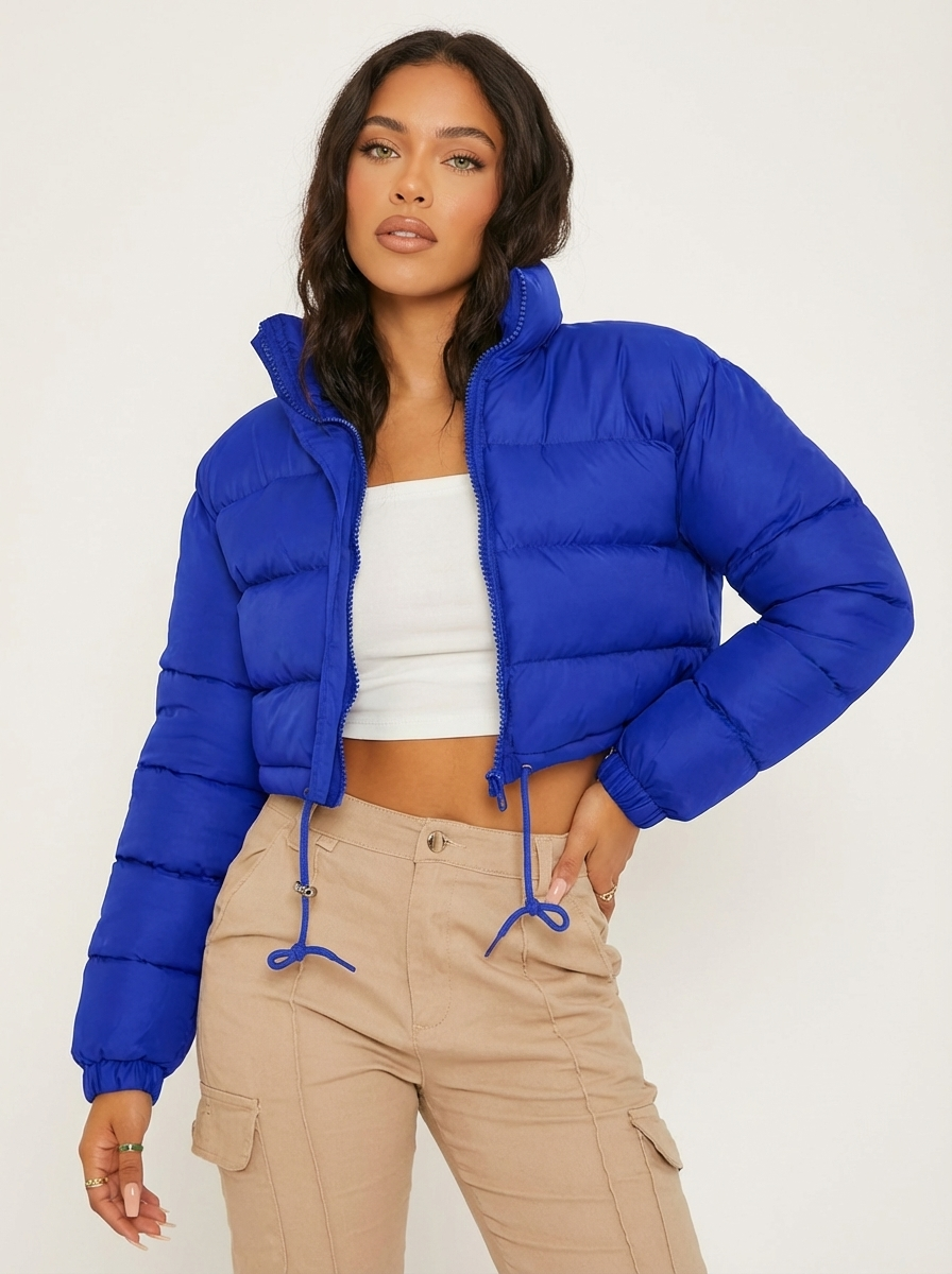 Blue - A woman wearing a dark brown cropped puffer jacket with an integrated hood, featuring horizontal quilting, a front zip closure, and adjustable drawstring toggles at the hem, paired with black high-waisted jeans.