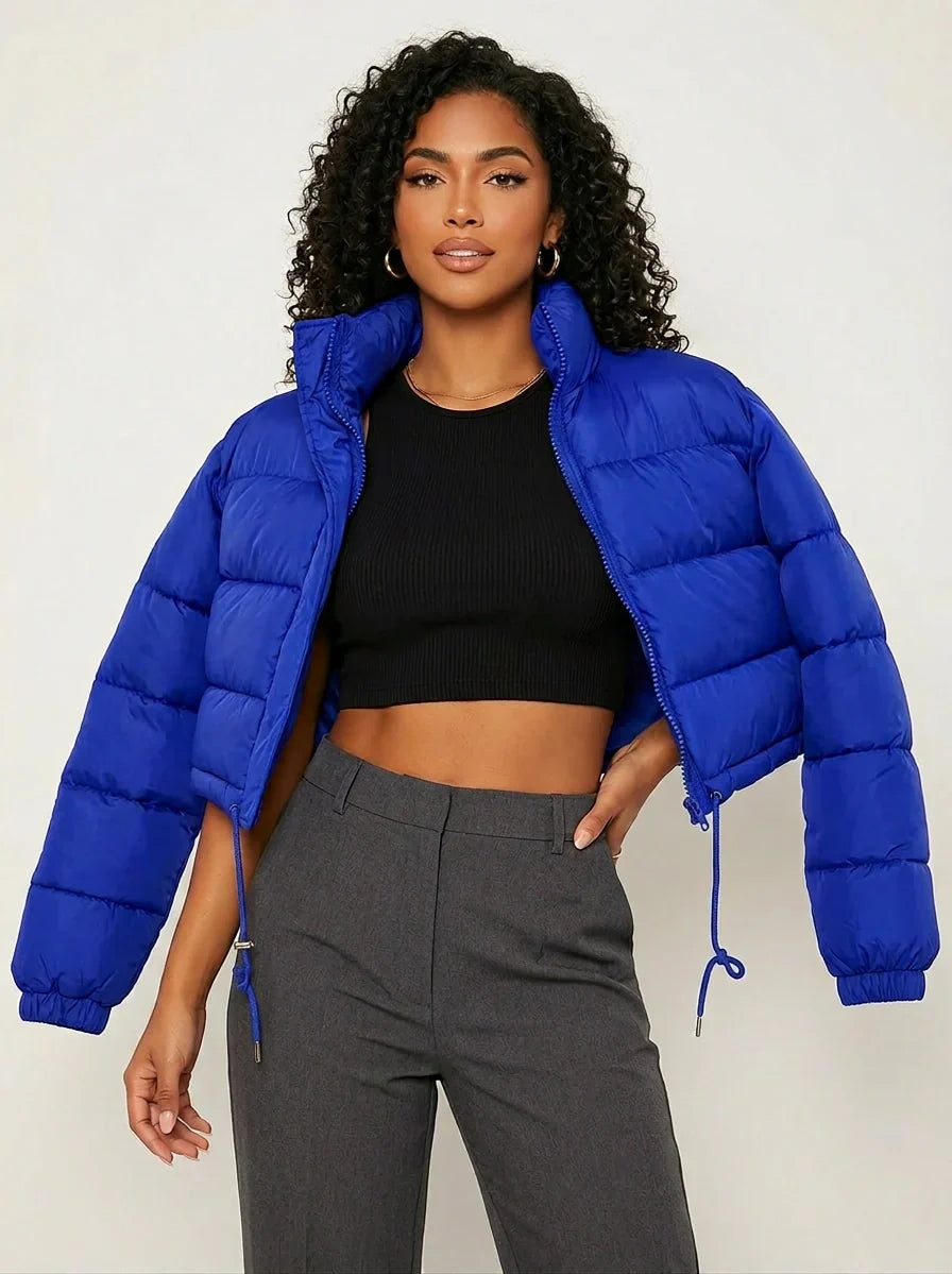 Blue - A woman wearing a dark brown cropped puffer jacket with an integrated hood, featuring horizontal quilting, a front zip closure, and adjustable drawstring toggles at the hem, paired with black high-waisted jeans.