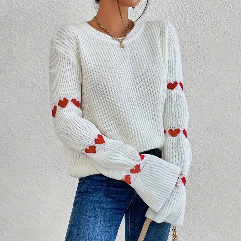 White - Women's white ribbed knit sweater featuring a crew neckline and long sleeves adorned with horizontal bands of red heart motifs, styled with blue jeans.