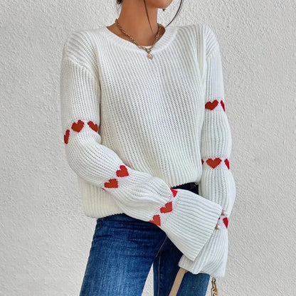White - Women's white ribbed knit sweater featuring a crew neckline and long sleeves adorned with horizontal bands of red heart motifs, styled with blue jeans.