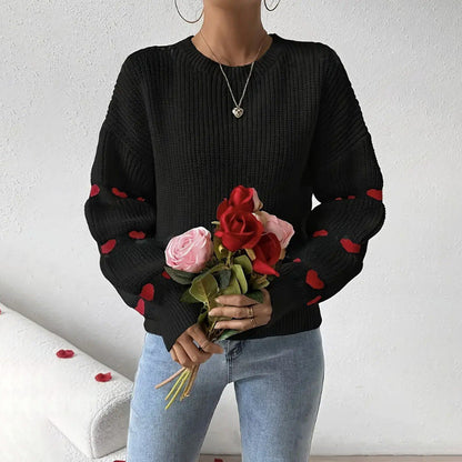 Black - Women's white ribbed knit sweater featuring a crew neckline and long sleeves adorned with horizontal bands of red heart motifs, styled with blue jeans.