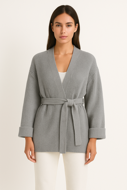 A woman wearing a light blue, ribbed knit wrap cardigan featuring a self-tie waist belt and wide folded cuffs, paired with cream trousers against a white background.