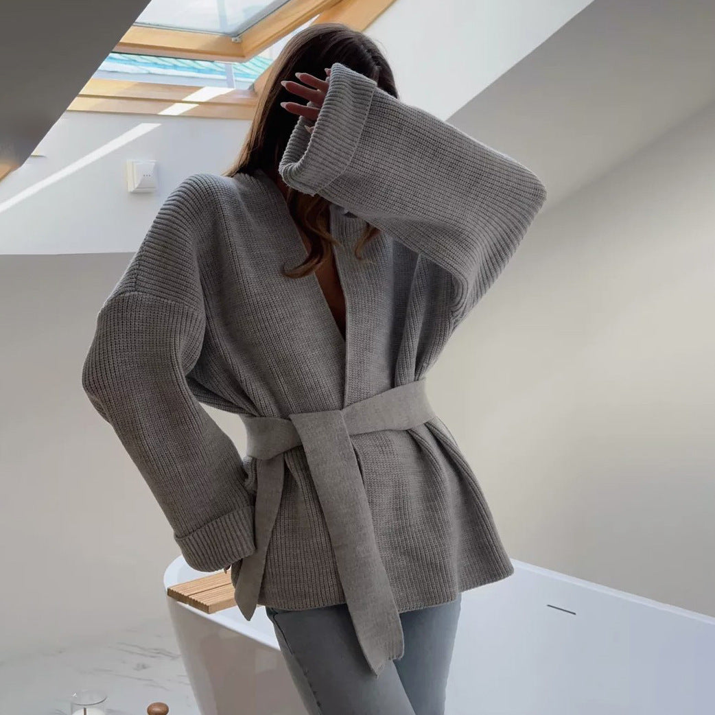 Grey - A woman wearing a light blue, ribbed knit wrap cardigan featuring a self-tie waist belt and wide folded cuffs, paired with cream trousers against a white background.
