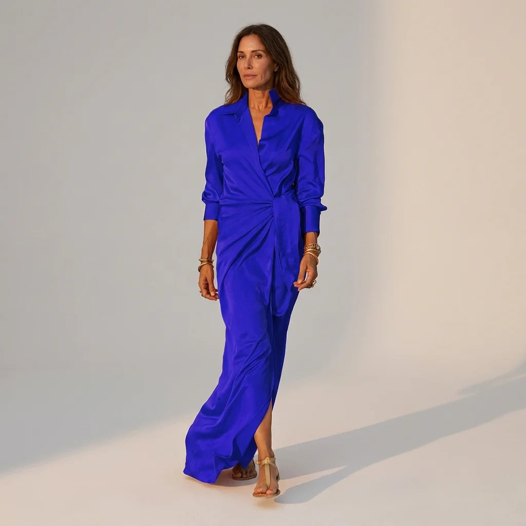 Blue - A woman wearing a long-sleeve, mustard-yellow satin wrap maxi dress with a collar, tied waist detail, and front split, posing against a neutral grey background.