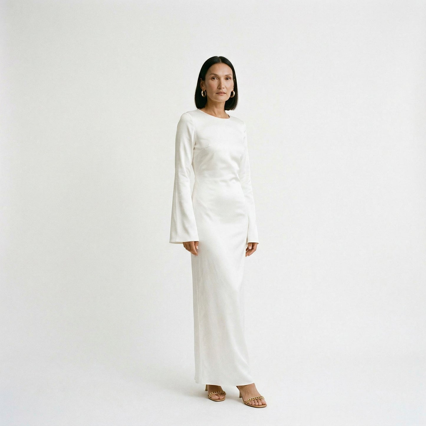 Cream - A woman wearing a long-sleeve chocolate brown satin maxi dress with a round neckline and flared cuffs, standing against a plain white background.