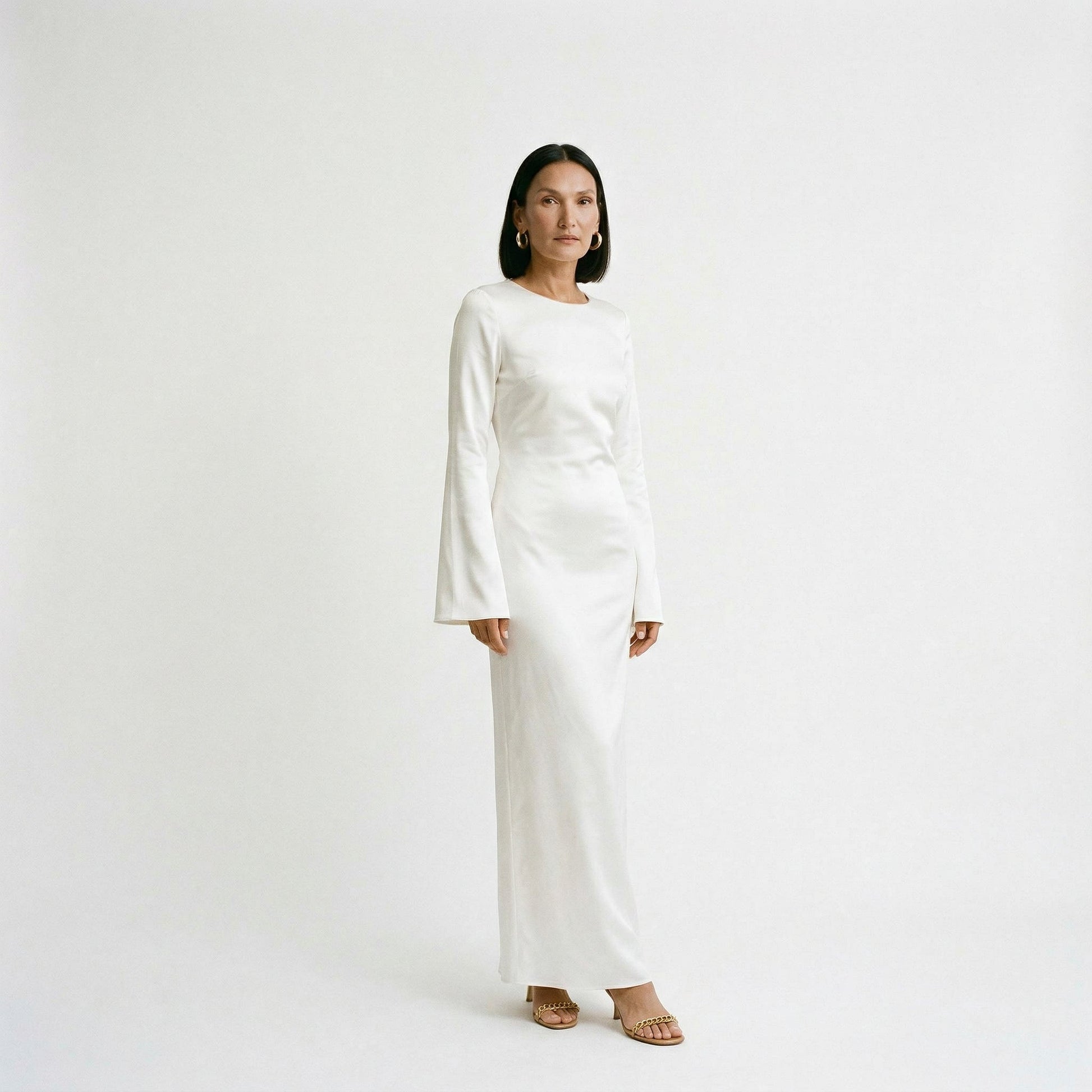 Cream - A woman wearing a long-sleeve chocolate brown satin maxi dress with a round neckline and flared cuffs, standing against a plain white background.