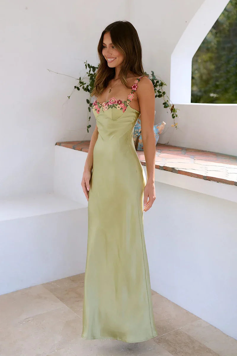 A woman wearing a sage green satin maxi dress with pink and red 3D floral appliqué along the scoop neckline and spaghetti straps, featuring a paneled bodice and a fluid drape.