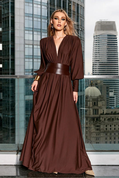 Brown - A woman wearing a chocolate brown satin maxi dress with a plunging V-neckline, ruched bodice, wide structured waist panel, and long bishop sleeves, standing against a city skyline background.